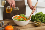 Woman adding olive oil into bowl with tasty salad at table in kitchen