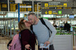 Couple of Caucasian travelers in front of the airport entrance. She gives him a kiss on the cheek