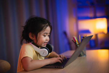 Wall Mural - Asian girl smiling, laughing, excited, looking, wearing white headphone and using laptop streaming night light in living room at home. Cute girl watching videos while tv Internet addiction concept.