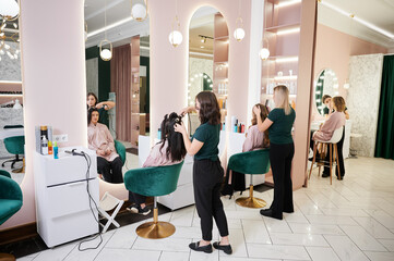 Wall Mural - Young women sitting in front of mirrors while hairdressers styling clients hair and makeup artist doing professional makeup. Makeup specialist and hairstylists working in modern beauty salon.