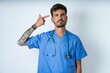 © Roquillo - Unhappy young caucasian doctor man wearing blue medical uniform makes suicide gesture and imitates gun with hand, curves lips keeps two fingers on temple, shoots, being tired of everything,