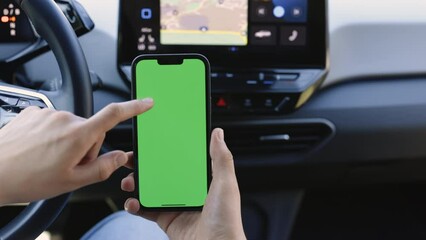 Man holding mobile phone on the background of a modern electric car interior. Green screen, blank phone, application. Swipe to the up down with your thumb. Driver in car uses a smartphone