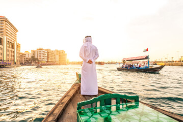 Wall Mural - Middle eastern man with traditional emirati kandura standing on abra boat and looking at horizon during sunset, concepts about travel and business