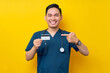 © Bangun Stock Photo - Portrait of a smiling cheerful young Asian male doctor or nurse wearing blue uniform pointing a finger at credit bank card isolated on yellow background. Healthcare medicine concept