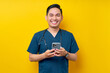 © Bangun Stock Photo - Smiling happy young Asian male doctor or nurse wearing blue uniform holding mobile phone and looking at a camera isolated on yellow background. Healthcare medicine concept