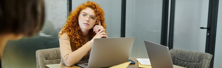 Wall Mural - serious redhead entrepreneur looking at blurred business partner near laptops in office, banner