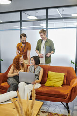 Wall Mural - positive businessmen near young women with laptop talking on couch in modern office, startup project