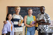 © AnnaStills - Group of four young successful intercultural students of highschool in casualwear standing in front of camera in classroom against blackboard