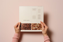  Woman's Hands In A Pink Sweater Holding A Coffee Menu Or Catalog With Coffee Beans, Top View In A Minimalist Setting