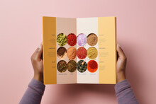 Female Hands Holding A Menu Or Catalog Featuring A Variety Of Spices, Shot From Above On A Pink Background