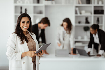 Wall Mural - Latin businesswoman ceo posing with digital tablet in office, smiling at camera during business meeting with colleagues