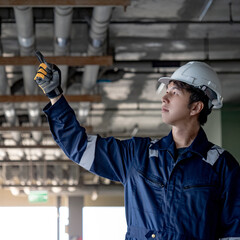 Portrait of Asian confident man construction worker in protective uniform suit and safety helmet pointing at piping system in the building. Male maintenance engineer working at construction site