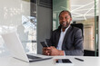 © Liubomir - Portrait of senior gray haired african american boss, man smiling and looking at camera at workplace inside office, businessman holding phone, using online app on smartphone.