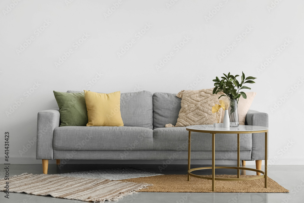 Cozy grey sofa with cushions and plant branches in vase on table near light wall