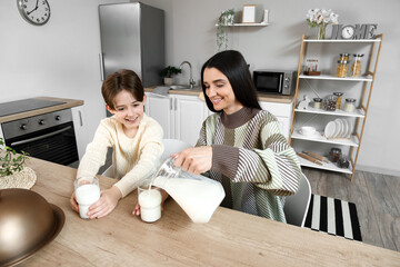 Poster - Happy little boy with his mother sitting at table and pouring fresh milk into glasses in kitchen