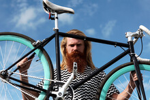 Portrait Of Middle-aged Man With Long Blond Hair And Full Red Beard Holding Retro Bike And Looking At Camera Against Blue Sky Background 