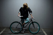 Young Bearded Man With Long Blond Hair Standing Beside His Retro Bike With Blue Rims In Underground Garage And Looking Down    