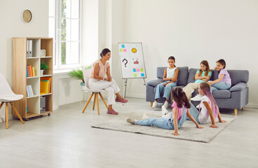 Wall Mural - Young smiling woman psychologist conducting mental health lesson for a group of school children girls sitting in a circle and having psychological training during therapy session in classroom.