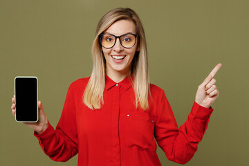 Wall Mural - Young surprised woman she wear red shirt casual clothes glasses hold in hand use mobile cell phone with blank screen workspace area pont index finger aside isolated on plain pastel green background