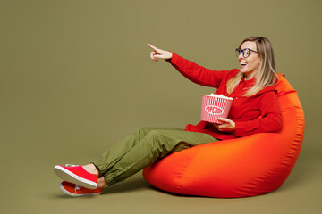 Wall Mural - Full body young woman wears red shirt casual clothes glasses sit in bag chair eat pop corn watch movie film point index finger aside on area isolated on plain pastel green background studio portrait.