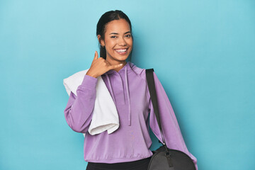 Wall Mural - Filipina with gym gear on blue studio showing a mobile phone call gesture with fingers.