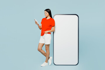 Full body side view young latin woman she wear orange red t-shirt casual clothes big huge blank screen mobile cell phone with area using smartphone isolated on plain pastel light blue cyan background.