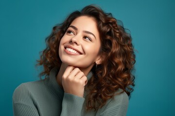 Wall Mural - Headshot portrait photography of a happy girl in her 20s putting the hand on the chin as if thinking against a teal blue background. With generative AI technology