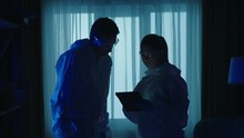 Male And Female Forensic Investigator Use A Tablet To List Evidence Found At A Crime Scene In A Dark Apartment. A Team Of Forensic Examiners, Illuminated By Blue Light From Police Sirens.