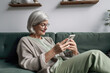 © Iryna - An elderly woman holds a smartphone and sits on a green sofa in the living room.