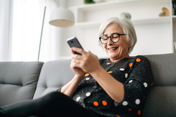 Modern lifestyle of the elderly. Stylish senior woman talking online using smartphone while relaxing on sofa.