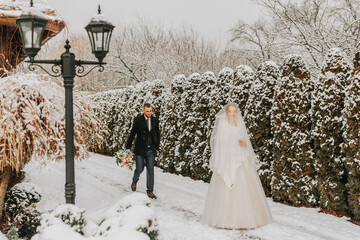 Wall Mural - Sensitive portrait of cheerful, beautiful newlyweds. The groom walks to his charming bride in the winter park. The bride in a wedding dress and poncho. The groom is dressed in a black coat