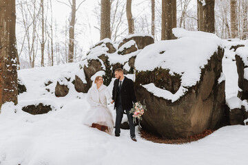 Wall Mural - Wedding in winter. Stylish bride in white mink coat and groom in black coat in forest with white snow in cold weather. The bride and groom are walking among rocks and large stones in the forest