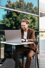 Red-haired businessman is typing on a laptop and working remotely outdoors. A busy male worker in a formal suit solves business problems against the backdrop of a building and writes an email.