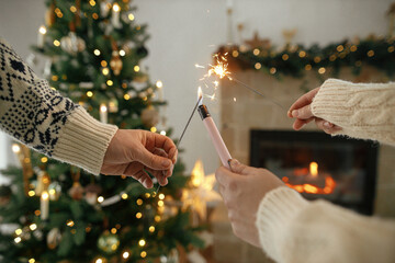 Wall Mural - Hands holding burning fireworks and lighter against modern fireplace and christmas tree with golden lights. Happy New Year! Friends and family celebrating with burning sparklers in hands