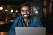 Generative AI Illustration Of Black Bearded Business Man Smiling Sand Looking At Camera While Sitting At Table In Front Of Laptop Against Dark Blurred Background With Lights
