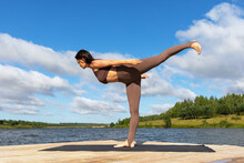 A Woman Leading A Healthy Lifestyle And Practicing Yoga Performs The Virabhadrasana Exercise, The Number Three Warrior Pose, Trains In Sportswear On The Shore Of The Lake On A Warm Sunny Morning
