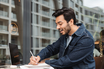 Positive Indian man, writing notes, using laptop computer