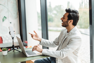 indian positive man in formal suit, gesturing hands and having video call while working