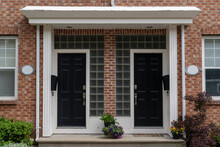 Two Black Metal Doors With Flower Pots On The Steps Of Duplex Houses. It Has A Red Brick Wall With White Wooden Trim. There Are Double Hung Windows On Both Sides Of The Building Entrance.