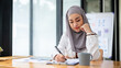 A focused Asian Muslim businesswoman is signing documents, or taking notes on paper at her desk