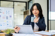 © bongkarn - A beautiful Asian businesswoman is working at her desk in a modern office, using a tablet