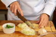 © Madu Oliveira - mãos de uma cozinheira usando Dolmã, preparando e picando alho e cebola sobre uma tabua de madeira e mais pote branco com salsa e cebolinha picados.