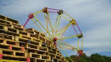 Revealing Big Wheel Roller Coaster Yellow And Red Colour Behind Stack Of Pallets Festival Park Scenery Partly Cloudy Summer Vibe Holiday Camping Trees In The Background
