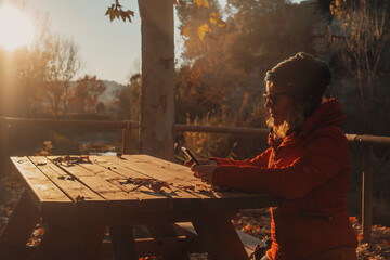 One woman with red jacket using mobile phone and outside wifi connection sitting on a bench and wooden table in the park during a golden autumn amazing sunset. Female people using cell device leisure