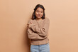 © wayhome.studio  - Pleased brunette Iranian woman daydreams embraces herself with both hands recalls romantic event keeps eyes closed smiles gently wears knitted jumper and jeans isolated over brown background.