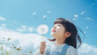 Photo of a little Asian girl blowing a dandelion, toddler child holding a dandelion flower on blue sky background, looking upwards, close-up summer portrait with copy space
