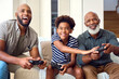 © Monkey Business - Multi-Generation Male Family Sitting On Sofa At Home Playing Video Game Together
