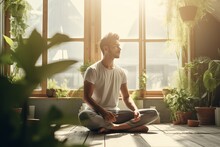 A Young Man In A Training Top T-shirt And Joggers Sitting In Yoga Asana Lotus Pose Meditating In A Sunlit Room With Green Plants