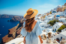 Woman In Blue Dress And Hat Looks Out Over The White Village Of Oia.