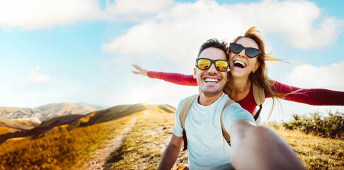 Wall Mural - Cheerful couple of hikers taking selfie on top of the mountain - Millennial guy and girl enjoying summertime day out laughing at camera together - Millenial travelers standing on nature background